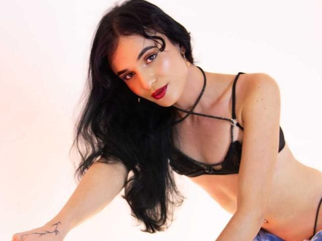 PamelaKander with long black hair, wearing a black strappy bra, showing her tits, red lipstick, and a tattoo on her right arm, leaning forward against a white background.
