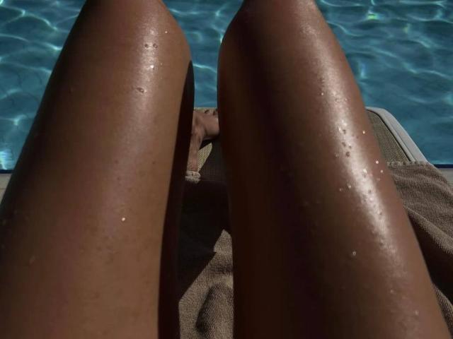 EricaLust's sun-kissed legs with water droplets, positioned by a pool, showing her smooth, dark brown skin and relaxed, tan-lined body, lying on a brown towel.