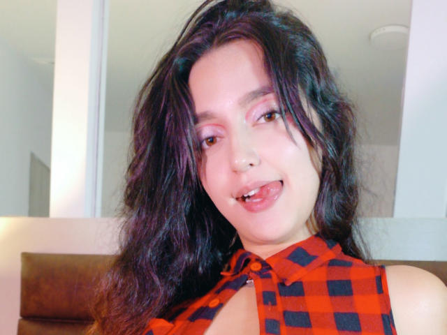 MelissaSmithers with wavy dark hair, pink eyeshadow, and a playful tongue-out expression, wearing a red and black checkered sleeveless top, showing a hint of her tits.