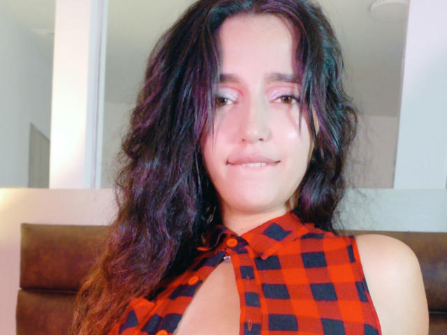 MelissaSmithers with wavy dark hair, pink eyeshadow, and a red-black checkered sleeveless top, showing cleavage, smiling gently. Her tits are partially visible through the top.