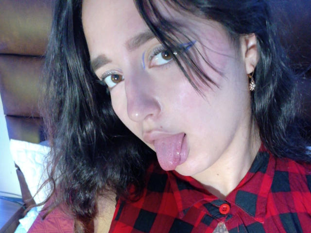 MelissaSmithers with wavy black hair, sticking out her tongue, wearing a red and black checkered shirt, brown eyes, and small earrings, has a slightly flushed face.