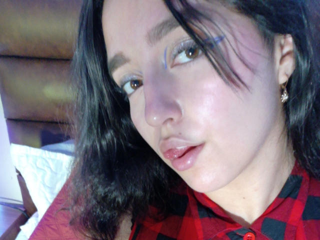 MelissaSmithers has shiny lips, dark wavy hair, and is wearing a red and black checkered shirt. She's looking to the side with light blue eyeliner.