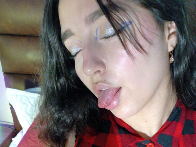 MelissaSmithers with closed eyes, glittery eyelids, blue facial markings, tongue out, black hair, red and black checkered shirt, and gold earrings, looking playful and slightly goofy.