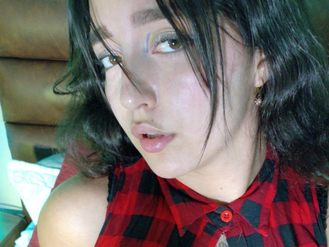 MelissaSmithers with shoulder-length black hair, wearing a red and black checkered shirt, has blue eyeshadow, small earrings, and slightly parted glossy lips, looking to the side.