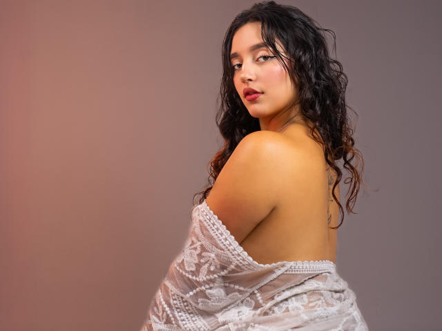 MelissaSmithers with curly black hair, looking over her shoulder, wearing a white lace off-shoulder top, exposing her bare back and ass. Her skin is tan, lips are red.