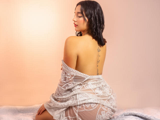 MelissaSmithers with dark wavy hair, sitting, back to camera, white lace dress slipping down, exposing a tattoo on her upper back, soft pink lighting, slightly parted red lips.