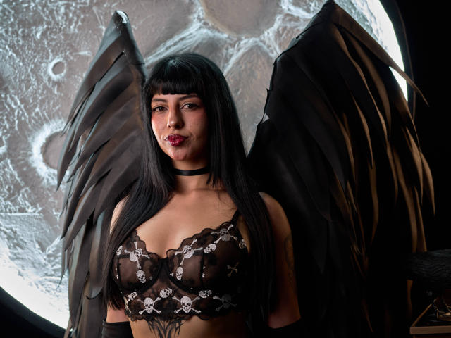 KatherinContin with black wings, black lace bra, and choker, showing her tits, long black hair, and red lipstick. Her wings are open, and she's looking at the camera.
