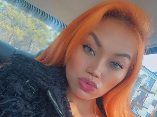 ForeignMaia with bright orange hair, full pink lips, green eyes, and a nose piercing, wearing a black textured top, looking at the camera with a pouty expression.