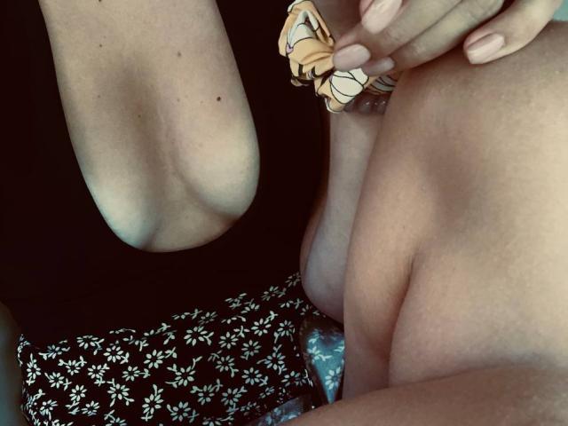 EricaLust's bare tits are visible, with her right hand holding her left arm, wearing floral shorts and a black top, and a patterned scrunchie on her wrist.