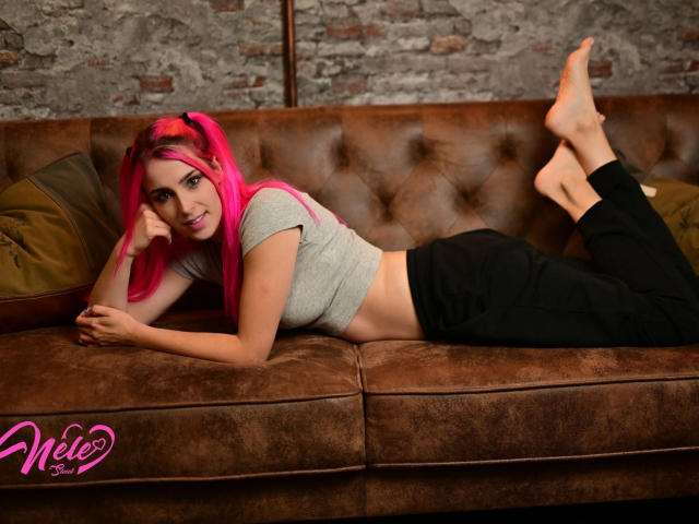 NeleSweet-hot with pink hair, lying on a brown couch, wearing a grey crop top and black pants, showing her ass and midriff, smiling at the camera.