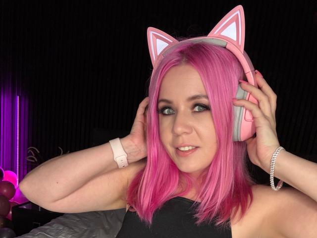 MayaMaibach-hot with pink hair, cat ears, and pink headphones, holding them on her ears, smiling, wearing a black top, pink watch, and silver bracelet.