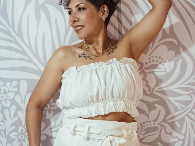 JuliaNewell with short curly hair, wearing a white strapless top and high-waisted pants, has tattoos on her chest, smiling, arm raised, wearing a gold necklace.