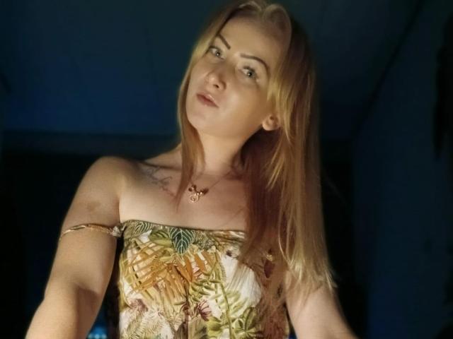 LeilahotX with blonde hair, wearing a strapless pineapple-patterned dress, slightly tilted head, and a gold necklace, looking off-camera with a subtle smile. Her tits are small.