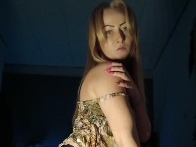 LeilahotX with blonde hair, staring down, hand on shoulder, wearing a snake-pattern dress, dark background, shadow on face, pink nails, looking serious, slightly turned, soft lighting.