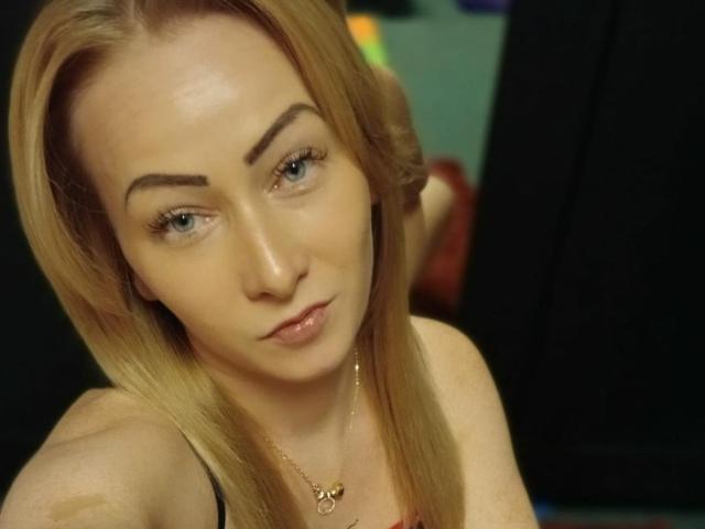 LeilahotX with blonde hair, light skin, and thick eyebrows, looking slightly to the side with a subtle, confident smile. She's wearing a necklace and has a relaxed expression.