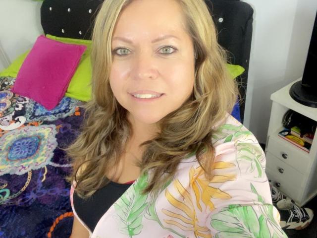 AlesanndraSexy has light brown wavy hair, green eyes, and a slight smile. She's wearing a floral pink shirt over a black top, with visible tits. Bedroom setting.