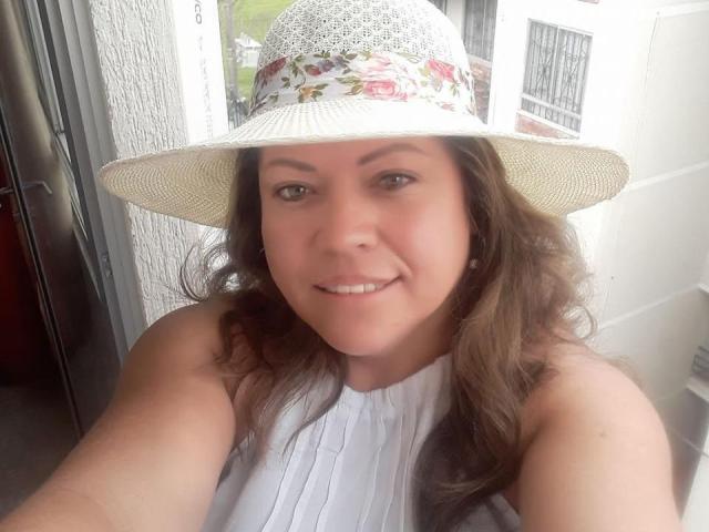 AlesanndraSexy with wavy brown hair, wearing a white floral sunhat and sleeveless white top, smiling at the camera. She has cute, small tits and is indoors.