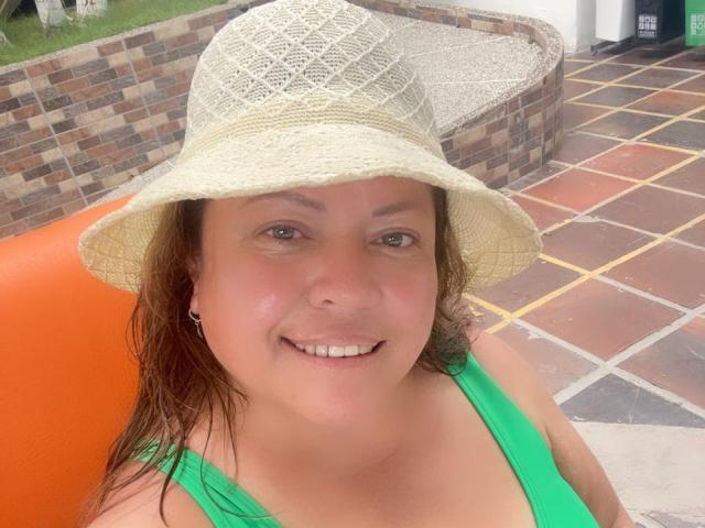 AlesanndraSexy with a straw hat, green tank top, and a smile, showing her tits in a photo. She's got wet brown hair and a cute face.