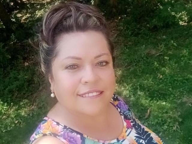 AlesanndraSexy has light skin, brown eyes, and a smile showing teeth. She's wearing a floral dress, pearl earrings, and has her brown hair in a messy updo.
