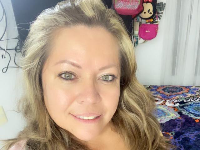 AlesanndraSexy has light green eyes, blonde wavy hair, and a slight smile. She's showing her white teeth, and her tits are not visible in the image.