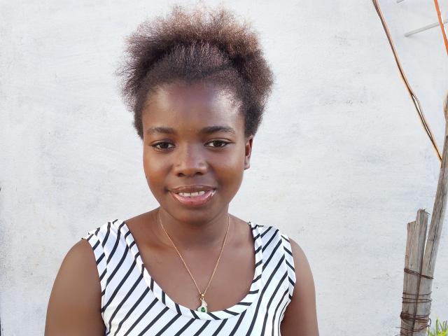 Fagilah has dark brown skin, short curly black hair, small nose, smiling with visible teeth, wearing a striped black and white sleeveless top, and a gold necklace.
