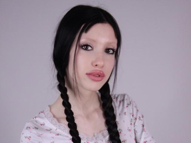 StellaBloum has pale skin, black braided hair, and pink lipstick. She's wearing a white floral shirt with lace. Her tits are small, and she's looking at the camera.