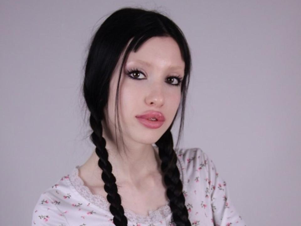 StellaBloum has pale skin, black braided hair, and pink lipstick. She's wearing a white floral shirt with lace. Her tits are small, and she's looking at the camera.