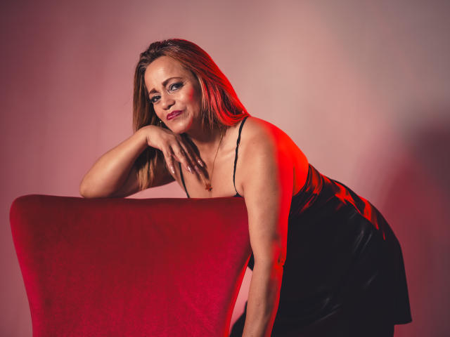 MaryMidori with red hair, black dress, leaning on red chair, smiling, hand on chin, red light, tits visible, attractive, confident, sexy.
