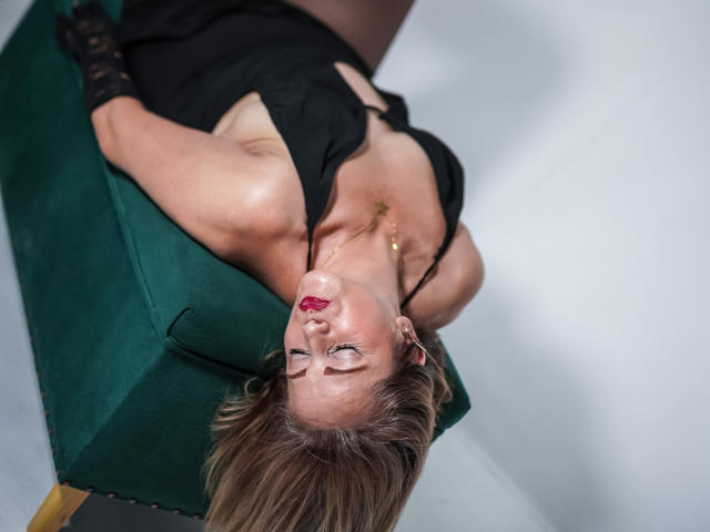 MaryMidori with blonde hair, red lipstick, black dress, and black gloves, leaning back on a green chair, eyes closed, head tilted, showing her tits and gold necklace.