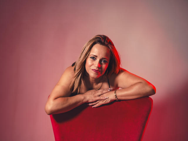 MaryMidori leans on a red chair, topless, with hands in front, displaying her tits and a slight smile, under red and pink lighting.