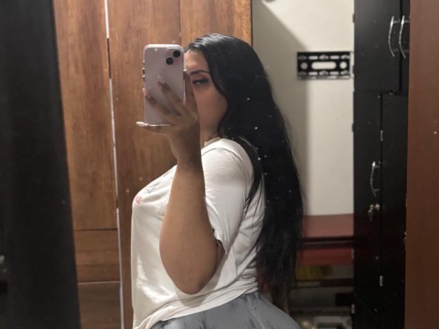 AmaliaDalton with long black hair, white shirt, and gray skirt, holding a pink phone, taking a mirror selfie, showing her curvy, big tits, and side butt.