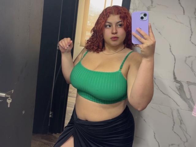 RoseDijon with curly red hair, green ribbed crop top, and black skirt, taking a mirror selfie, holding a purple phone, showing her tits and a thigh cutout.