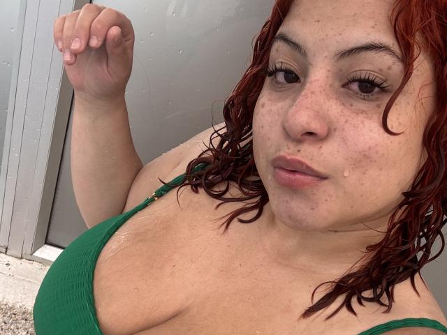 RoseDijon with wet red curly hair, green top, showing her tits, freckled face, and subtle smile, holding up her right hand, water droplets on her skin.