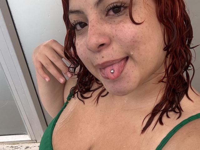 RoseDijon has wet red hair, freckles, and is sticking out her tongue with a tongue piercing. She's wearing a green top, and water droplets are on her chest.