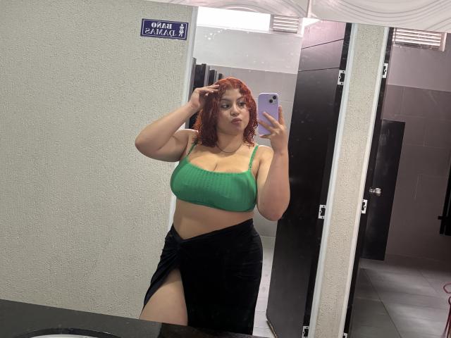 RoseDijon with curly red hair, green bra showing her big boobs, black skirt with a thigh gap, holding a phone in a bathroom mirror selfie.