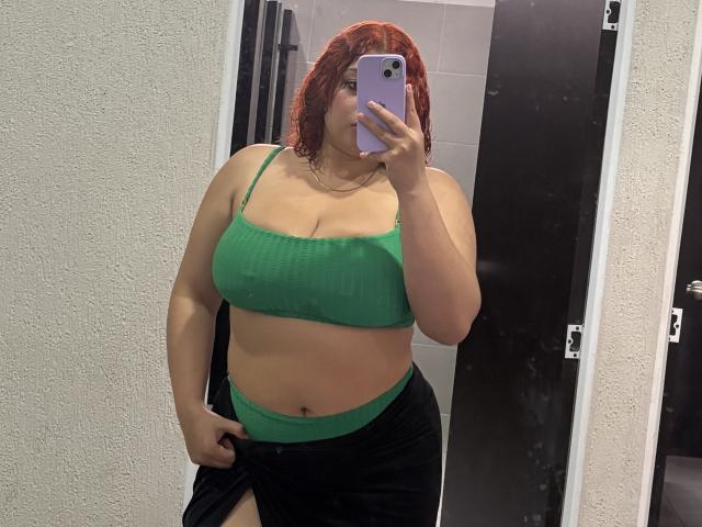 RoseDijon with red hair, green ribbed crop top showing her tits, black skirt with green waistband, holding phone in mirror selfie, hand on skirt, in bathroom.