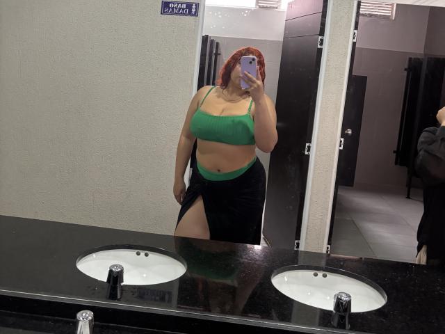 RoseDijon with red hair, green sports bra, showing her tits and a black skirt with a thigh slit, taking a mirror selfie in a bathroom.