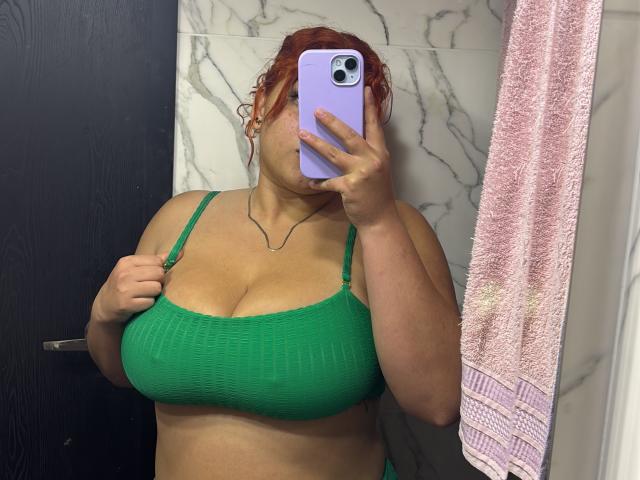 RoseDijon with red curly hair, green ribbed bra, holding phone covering face, showing large boobs, green strap, pink towel on right, marble wall, black door, in bathroom.