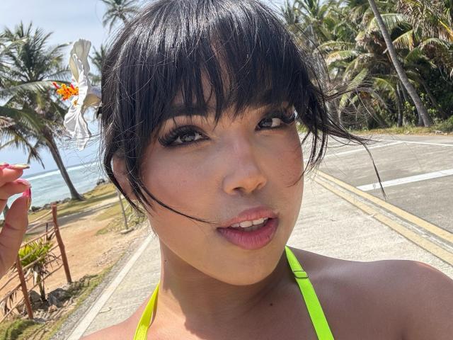 Heyly with wet black bangs, looking directly at camera, mouth slightly open, wearing neon green bikini top, tropical palm trees background.