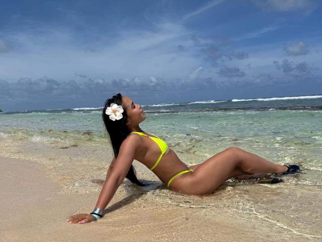 Heyly, with dark skin and a white flower in her hair, leans back in shallow water, wearing a yellow bikini, footjob.