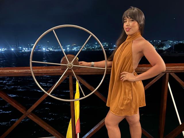 Heyly with big tits, sleeveless orange dress, leaning on shipwheel, hand on hip, city lights in dark night sky background.
