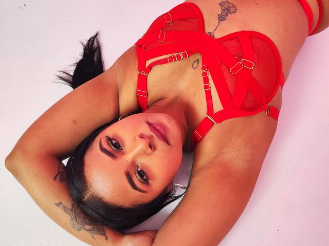 LunaaWilson with black hair, red strappy lingerie, and tattoos, lying on her back, arms above her head, looking at the camera with a slight smile. Boobs, red bra, tattoos.