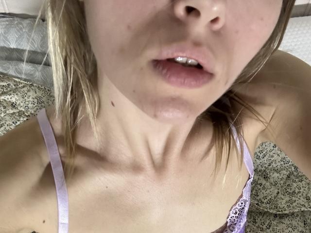 Das Gesicht von KrissWild mit leicht geöffneten, rosafarbenen Lippen, einem kleinen Muttermal auf der linken Seite ihres Halses und blonden Haaren, sie trägt einen lila Spitzen-BH, aufgenommen in einer engen Aufnahme von oben..