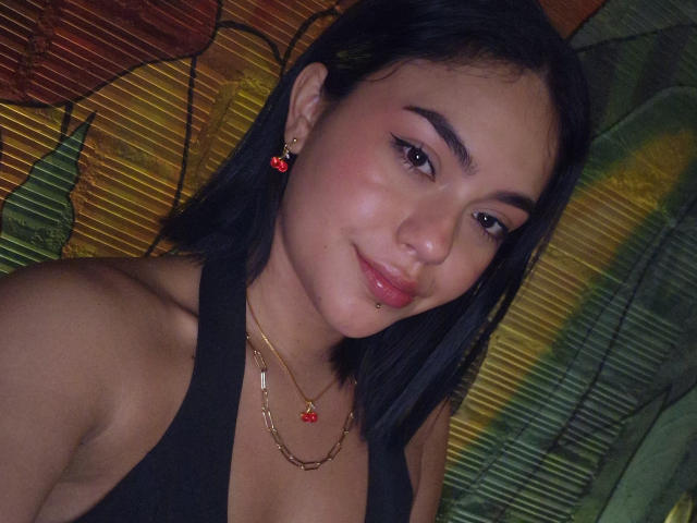 Vale69 has tan skin, black hair, and is smiling with pink lips. She's wearing a black tank top, gold chain with red flower, and red earrings. Boobs visible.