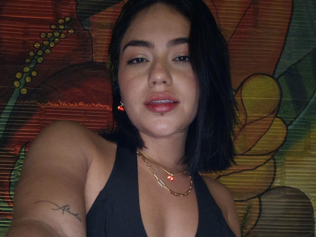 Vale69 has black hair, a gold necklace, red earrings, a lip piercing, and a tattoo on her right arm. She's wearing a black top, showing her tits.