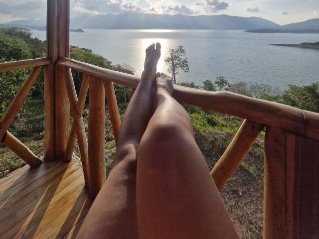 Vale69's legs stretched out on a wooden balcony, sun shining on their bare, smooth, brown skin, with their toes pointing towards a bright, reflective lake in the background.
