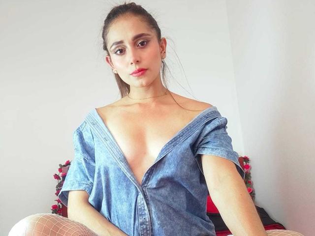 EliBaker with a serious face, wearing an off-shoulder blue denim shirt that reveals her tits, and white fishnet stockings, sitting on a bed with pink flowers.