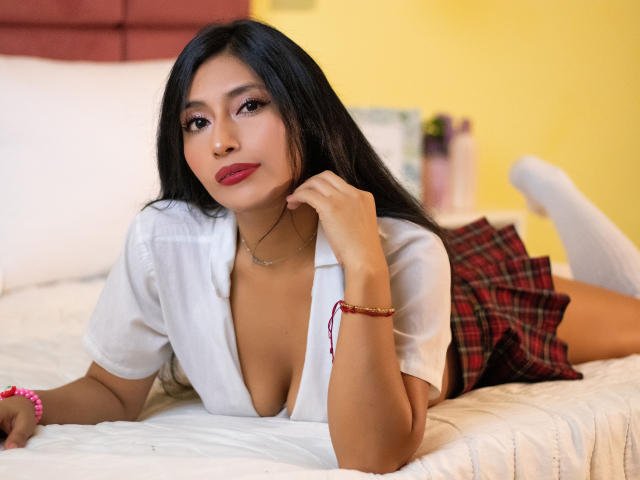 LucianaHernandez with long black hair, red lipstick, and a white shirt open showing her tits, lying on a bed in a red plaid skirt and white socks.