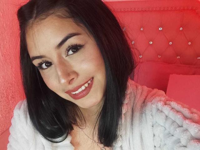 EmilyVv has black hair, brown eyes, and a cute smile with red lipstick. She's wearing a fluffy white robe, and the red background has a tufted, buttoned-up headboard.
