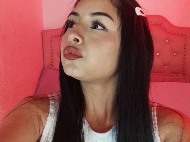EmilyVv with pouty lips, long black hair, white tank top, pink hair clip, and red-tinted skin, looking up with big eyes and long eyelashes.